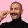 Smiling man with a mustache twirling the tip, wearing a black turtleneck against a pink background.